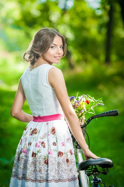 Beautiful girl wearing a nice white dress having fun in park with bicycle. Healthy outdoor lifestyle concept. Vintage scenery. Pretty blonde girl with retro look with bike and basket with flowers — Stock Photo, Image