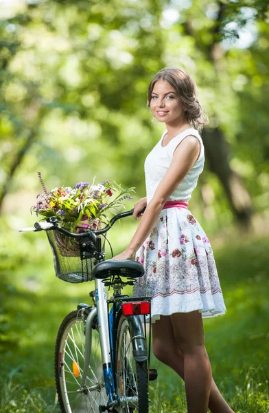 Beautiful girl wearing a nice white dress having fun in park with bicycle. Healthy outdoor lifestyle concept. Vintage scenery. Pretty blonde girl with retro look with bike and basket with flowers — Stock Photo, Image