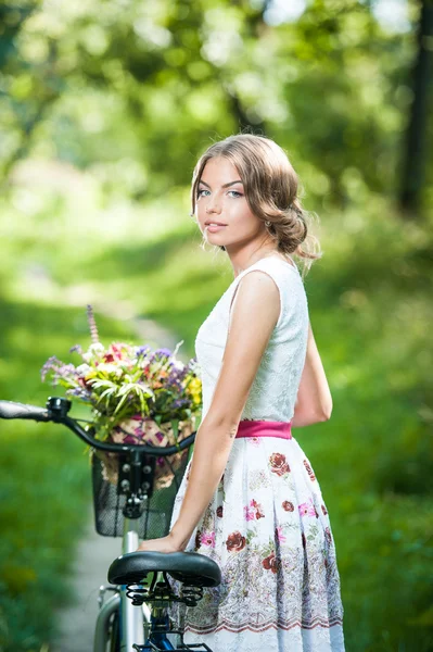 Krásná dívka, která nosí pěkné bílé šaty, baví se v parku s kol. venkovní zdravého koncept. Vintage scenérie. docela blondýna s retro vzhled s kolo a košík s květinami — Stock fotografie