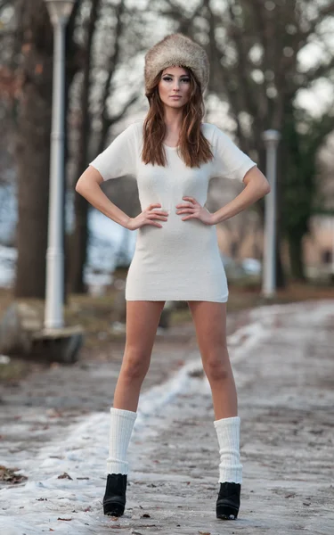 Портрет молодой красивой женщины, съемка на открытом воздухе в зимнем пейзаже. Чувственная брюнетка в коротком белом платье и меховой шапке позирует в парке, покрытом снегом. Модная женщина в холодный день — стоковое фото