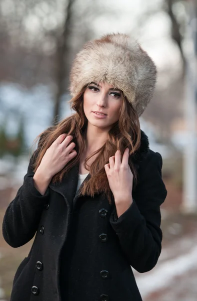 Retrato de mujer hermosa joven, tiro al aire libre en el paisaje de invierno. Chica morena sensual con abrigo y gorra de piel posando en un parque cubierto de nieve. La hembra a la moda en el día frío . —  Fotos de Stock