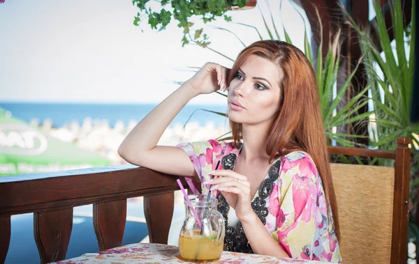 Attractive red hair young woman with bright colored blouse drinking lemonade on a terrace having blue sea in background. Gorgeous redhead model drinking fresh drink with straw in a summer day — Stock Photo, Image