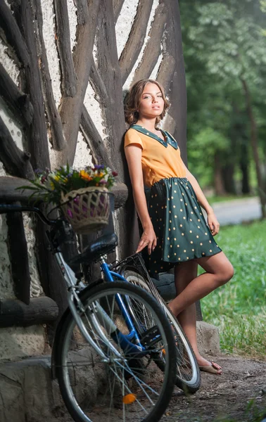 Vacker flicka klädd i en snygg klänning med college utseende ha kul i parken med cykel bära en vacker korg. Vintage landskap. Söt retro blonda tjejen för cykel och korg med blommor — Stockfoto