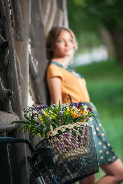 Vacker flicka klädd i en snygg klänning med college utseende ha kul i parken med cykel bära en vacker korg. Vintage landskap. Söt retro blonda tjejen för cykel och korg med blommor — Stockfoto