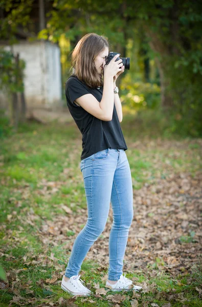 Muchacha joven atractiva tomando fotos al aire libre. Linda adolescente en jeans azules y camiseta negra tomando fotos en el parque otoñal. Retrato de otoño al aire libre de bastante adolescente divirtiéndose en el parque con cámara —  Fotos de Stock