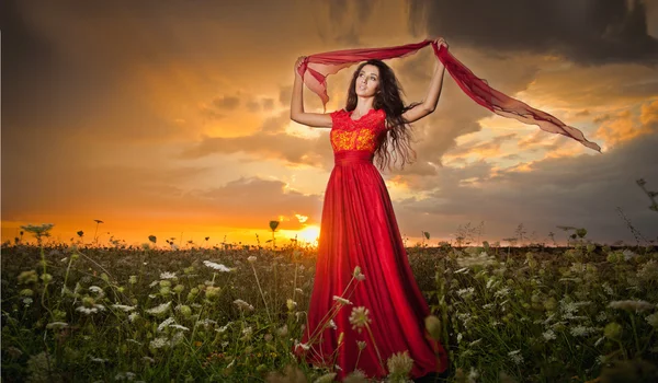 Belle jeune femme à la mode en robe rouge longue posant en plein air avec ciel nuageux dramatique en arrière-plan. Jolie fille brune aux cheveux longs avec une élégante robe de luxe, coup de soleil couchant . — Photo