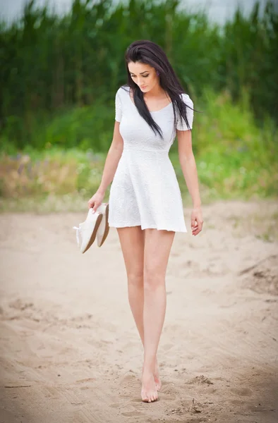 Chica morena atractiva con vestido blanco corto paseando descalzo en el camino del campo. Mujer hermosa joven caminando con zapatos en la mano con el bosque en el fondo. Mujer con piernas largas al aire libre — Foto de Stock