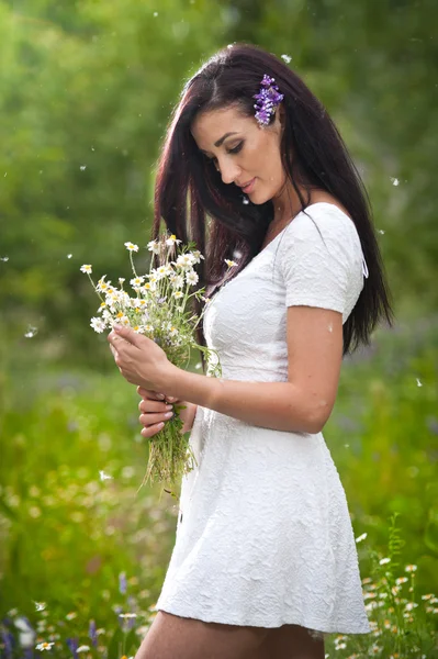 Joven mujer morena hermosa sosteniendo un ramo de flores silvestres en un día soleado. Retrato de atractiva hembra de pelo largo en vestido blanco, tiro al aire libre. Vista lateral de linda chica disfrutando de la naturaleza en verano — Foto de Stock