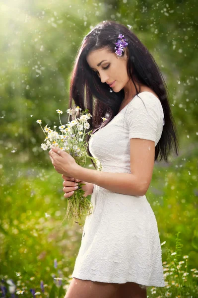 Young beautiful brunette woman holding a wild flowers bouquet in a sunny day. Portrait of attractive long hair female in white dress, outdoor shot. Side view of cute girl enjoying the nature in summer — Stock Photo, Image