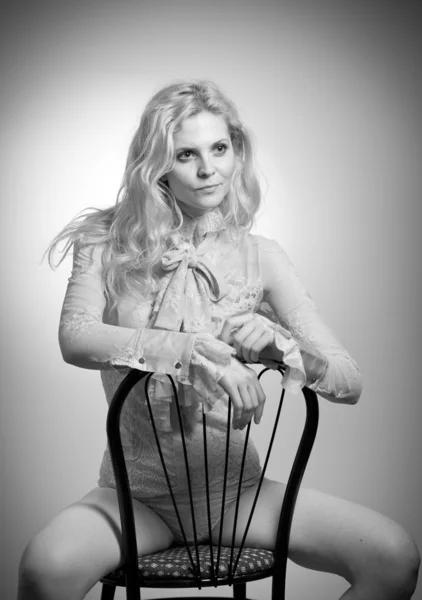 Attractive fair hair model with in elegant nude blouse sitting provocatively on chair, studio shot. Fashion portrait of a sensual blonde woman in classic blouse with long sleeves and ribbon on chair — Stock Photo, Image