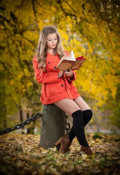 Piękna dziewczynka elegancki z pomarańczowy płaszcz czytanie, siedząc na pniu jesienny park. Młode ładne kobiety z blond włosami spędzać czas w upadku. Długie nogi zmysłowa blondynka relaks z książką w lesie — Zdjęcie stockowe