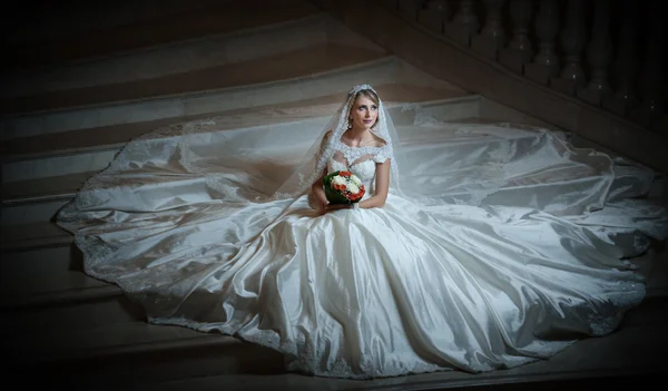 Young beautiful luxurious woman in wedding dress sitting on stair steps in semi-darkness. Bride with huge wedding dress holding her bouquet. Seductive blonde bride with gorgeous gown posing — Stock Photo, Image