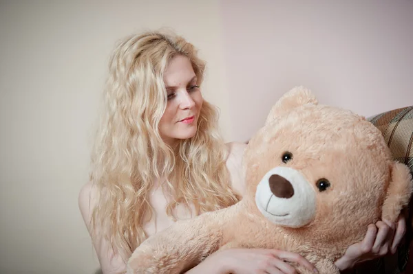 Young blond sensual woman sitting on sofa relaxing with a huge teddy bear. Beautiful girl with comfortable clothes relaxing on the couch with a toy. Attractive blonde in cozy scenery indoor — Stock Photo, Image