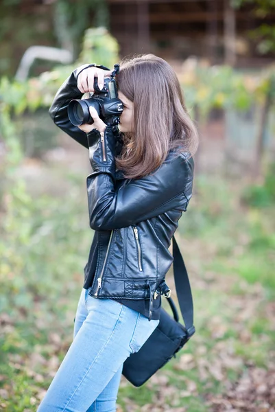 Atrakcyjny dziewczynka fotografowanie na zewnątrz. Cute nastolatka w niebieskie jeansy i czarną skórzaną kurtkę robienia zdjęć w jesienny park. Zewnątrz portret ładna nastolatka zabawy w parku z aparatu — Zdjęcie stockowe