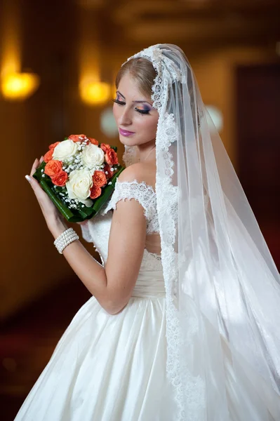 Jeune belle femme luxueuse en robe de mariée posant dans un intérieur luxueux. Mariée avec un long voile tenant son bouquet de mariage. Mariée blonde séduisante avec robe à la mode, plan intérieur — Photo