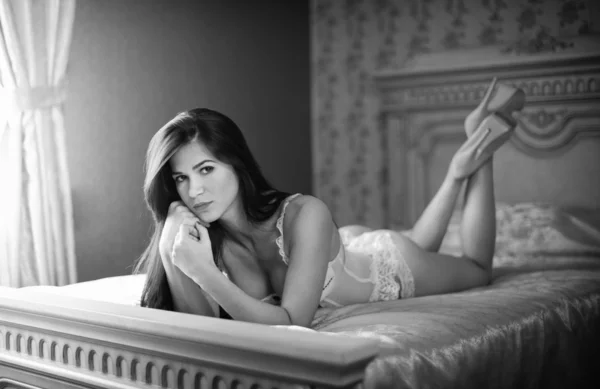 美丽的性感黑发年轻女人穿着白色睡衣躺在床上。与挑衅，构成室内射击的身体吸引力的时尚女性。感性的女孩，与在酒店床上的高跟鞋 — 图库照片