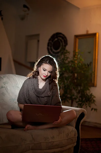 Beautiful young woman sitting on sofa working on laptop having a red gramophone near her, in boudoir scenery. Attractive brunette girl with long hair and long legs laying down on couch with a laptop — Stock Photo, Image