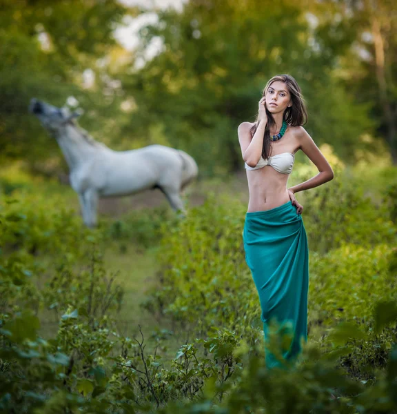 Giovani donne in gonna lunga blu e reggiseno bianco al tramonto nella foresta con un cavallo bianco sullo sfondo. Bella giovane donna con i capelli lunghi in giardino con cavallo selvatico. Ragazza e cavallo nel campo — Foto Stock