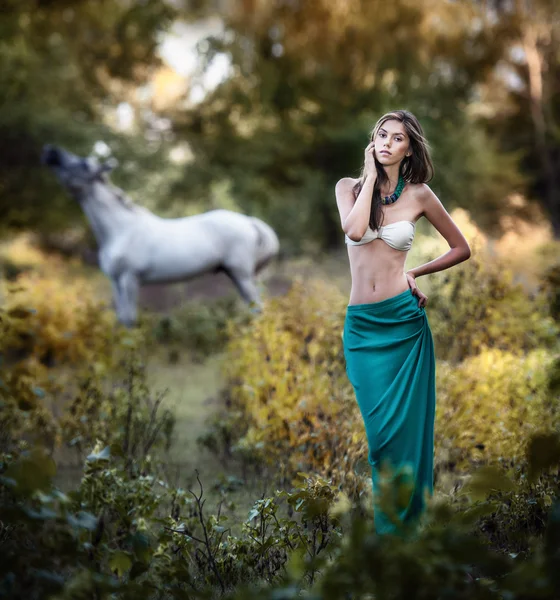 Junge Frauen in einem blauen langen Rock und weißen BH bei Sonnenuntergang im Wald mit einem weißen Pferd im Hintergrund. Schöne junge Frau mit langen Haaren im Garten mit Wildpferd. Mädchen und Pferd auf dem Feld — Stockfoto