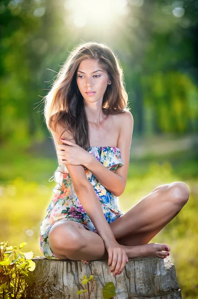 Young beautiful long hair woman wearing a multicolored dress posing on a stump in a green forest. Fashionable sexy attractive girl sitting on hub in a sunny day. Gorgeous girl in garden in summer day. — Stock Photo, Image