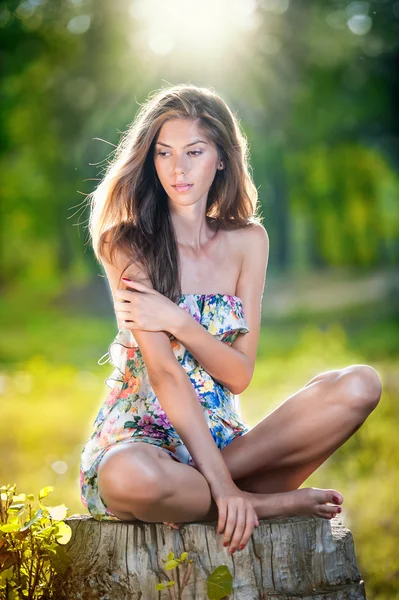 Jeune belle femme aux cheveux longs portant une robe multicolore posant sur une souche dans une forêt verte. Fille sexy attrayant à la mode assis sur le moyeu dans une journée ensoleillée. Superbe fille dans le jardin en journée d'été . — Photo