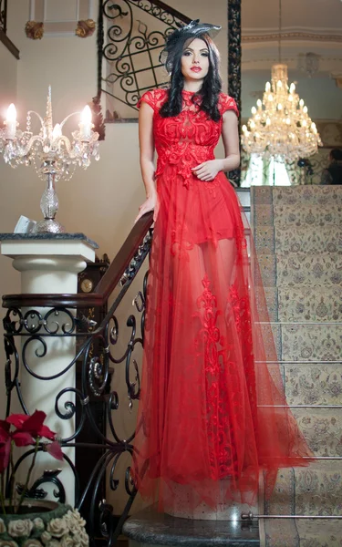 Het mooie meisje in een lange rode jurk poseren in een vintage scène. jonge mooie vrouw draagt een rode jurk in een oud hotel. sensuele elegante jonge vrouw in rode lange jurk binnen schot. — Stockfoto