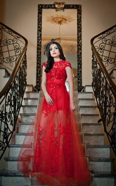 The beautiful girl in a long red dress posing in a vintage scene. Young beautiful woman wearing a red dress in an old hotel. Sensual elegant young woman in red long dress indoor shot. — Stock Photo, Image