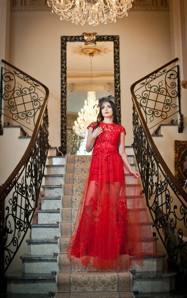 The beautiful girl in a long red dress posing in a vintage scene. Young beautiful woman wearing a red dress in an old hotel. Sensual elegant young woman in red long dress indoor shot. — Stock Photo, Image