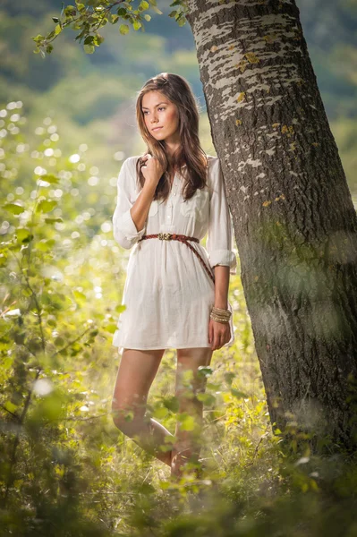 Atractiva joven en vestido corto blanco posando cerca de un árbol en un día soleado de verano. Hermosa chica disfrutando de la naturaleza en un bosque verde. Retrato de sensual hembra en blanco soñando despierto en un prado —  Fotos de Stock