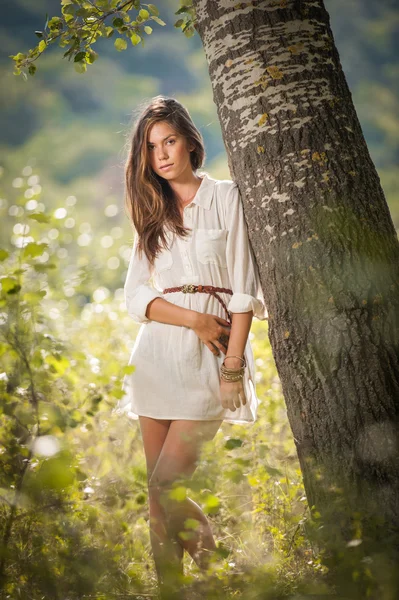 Jovem atraente em vestido curto branco posando perto de uma árvore em um dia ensolarado de verão. Menina bonita apreciando a natureza em uma floresta verde. Retrato de mulher sensual em branco sonhando acordado em um prado — Fotografia de Stock