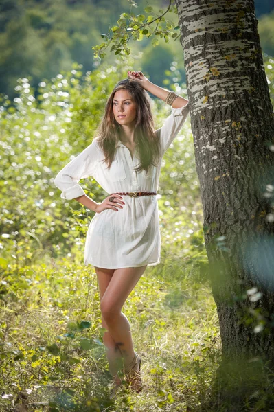 Привлекательная молодая женщина в белом коротком платье позирует возле дерева в солнечный летний день. Красивая девушка наслаждается природой в зеленом лесу. Портрет чувственной женщины в белом дневном сне на лугу — стоковое фото