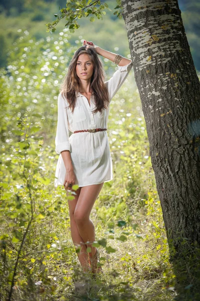 Attractive young woman in white short dress posing near a tree in a sunny summer day. Beautiful girl enjoying the nature in a green forest. Portrait of sensual female in white daydreaming in a meadow — Stock Photo, Image