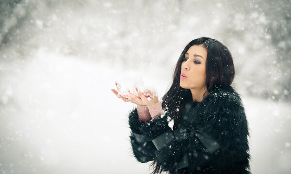 Woman blowing snow from her hands enjoying the winter. Happy brunette girl playing with snow in the winter landscape. Beautiful young female on winter background