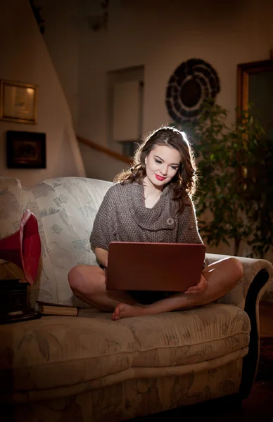 Beautiful young woman sitting on sofa working on laptop having a red gramophone near her, in boudoir scenery. Attractive brunette girl with long hair and long legs laying down on couch with a laptop — Stock Photo, Image