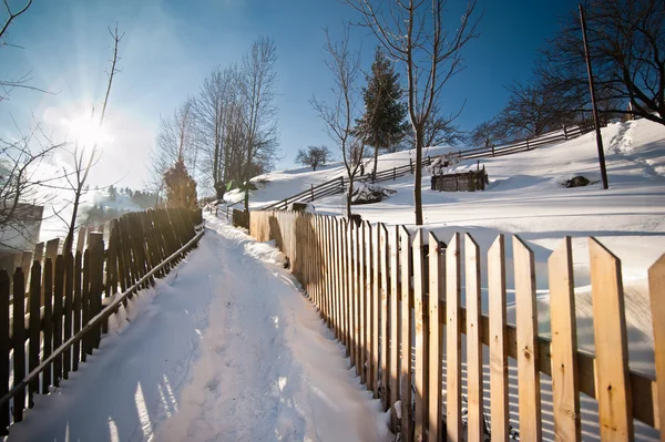 Carretera estrecha cubierta de nieve en el campo. Paisaje invernal con árboles nevados, carretera y valla de madera. Día frío de invierno en el campo. Montañas tradicionales Cárpatos paisaje del pueblo, Rumania — Foto de Stock