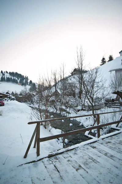 Ponte de madeira em uma aldeia romena tradicional através de um pequeno rio. Ponte sobre rio congelado. Inverno paisagem rural, Moeciu, Roménia. Ponte de madeira sobre pequeno riacho congelado coberto de neve — Fotografia de Stock
