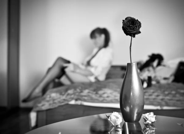 Таинственная брюнетка кладет свой чулок с цветами и вазой на переднем плане. Чувственная женщина, одевающаяся, сидящая на кровати и ваза с розой на столе. Крытый снимок, черно-белое фото . — стоковое фото