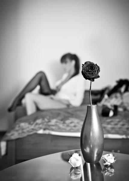 Mysterious brunette putting her stocking with flowers and vase foreground. Sensual woman dressing up sitting on bed and a vase with a rose on a table. Indoor shot, black and white photo. — Stock Photo, Image