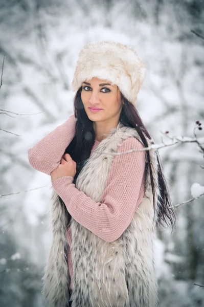 Женщина в белом меховом колпаке и овчины улыбается, наслаждаясь зимними пейзажами в лесу. Вид сбоку на счастливую брюнетку, позирующую в зимнем пейзаже. Красивая молодая женщина на зимнем фоне — стоковое фото
