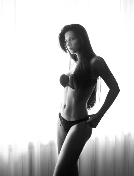 Jovem bela mulher sexy em lingerie preta posando na luz da janela perto de cortinas. Sensual morena longo cabelo feminino com corpo perfeito ficar perto de uma janela. Menina atraente, foto em preto e branco — Fotografia de Stock