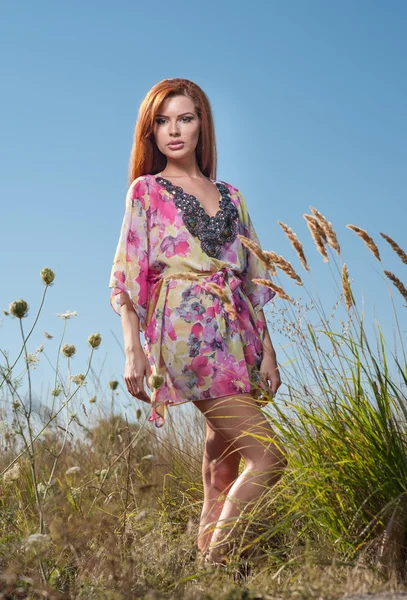 Mulher bonita no campo de flores selvagens no fundo do céu azul. Retrato de atraente menina de cabelo vermelho com cabelos longos relaxante na natureza, tiro ao ar livre. Senhora em vestido multicolorido apreciando a natureza — Fotografia de Stock