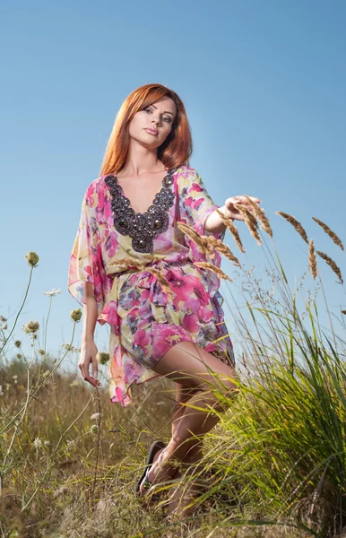 Mooie jonge vrouw in wilde bloemen veld op blauwe hemelachtergrond. Portret van aantrekkelijke rood haar meisje met lange haren ontspannen in de natuur, buiten schot. Lady in veelkleurige jurk genieten van natuur — Stockfoto
