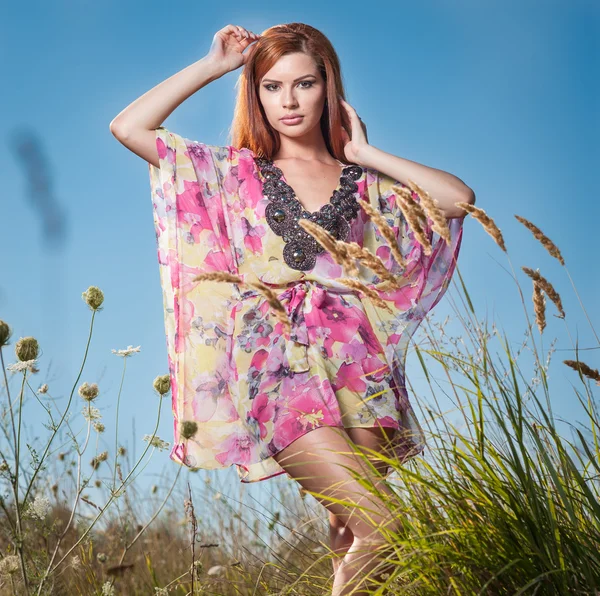 Mooie jonge vrouw in wilde bloemen veld op blauwe hemelachtergrond. Portret van aantrekkelijke rood haar meisje met lange haren ontspannen in de natuur, buiten schot. Lady in veelkleurige jurk genieten van natuur — Stockfoto