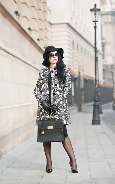 Atractiva joven en una foto de moda de invierno. Hermosa chica joven de moda en negro posando en la avenida. Elegante morena con sombrero, gafas de sol y bolso en el paisaje urbano . — Foto de Stock