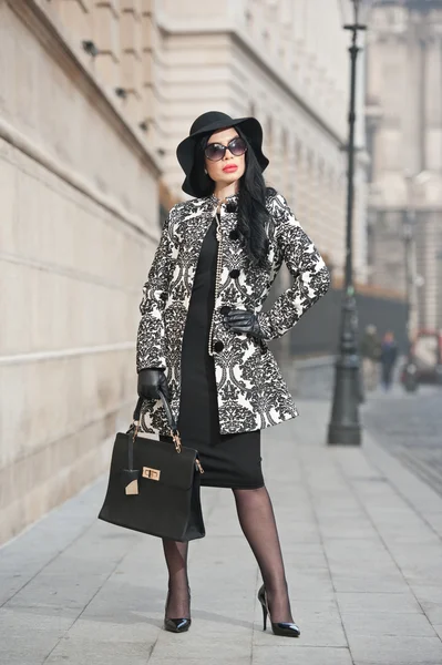 Attractive young woman in a winter fashion shot. Beautiful fashionable young girl in black  posing on avenue. Elegant brunette with hat, sunglasses and handbag in urban scenery. — Stock Photo, Image