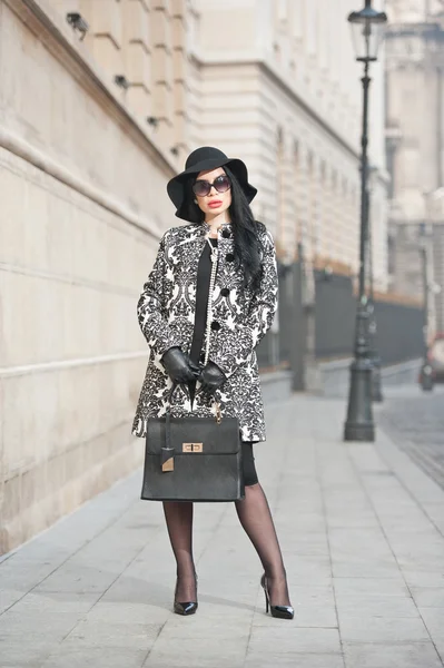 Atractiva joven en una foto de moda de invierno. Hermosa chica joven de moda en negro posando en la avenida. Elegante morena con sombrero, gafas de sol y bolso en el paisaje urbano . — Foto de Stock