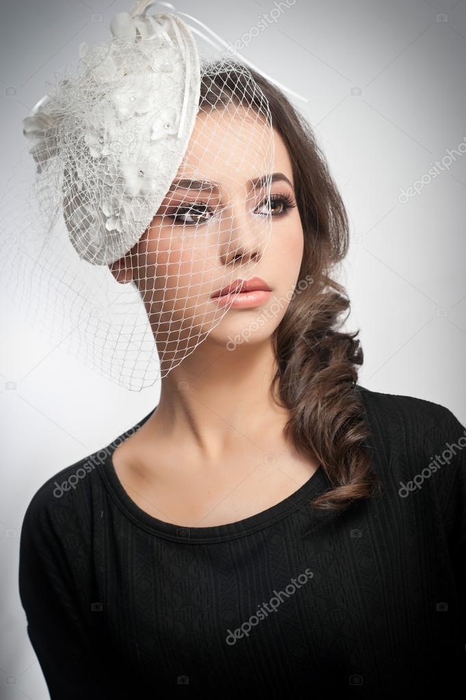 Hairstyle and make up - beautiful young girl art portrait. Cute brunette with white cap and veil, studio shot. Attractive female with beautiful lips and eyes in black blouse, over white