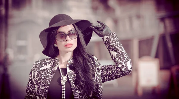 Atractiva joven en una foto de moda de invierno. Hermosa chica joven de moda en negro posando en la avenida. Elegante morena con sombrero, gafas de sol y guantes en el paisaje urbano . — Foto de Stock