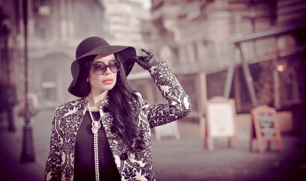 Atractiva joven en una foto de moda de invierno. Hermosa chica joven de moda en negro posando en la avenida. Elegante morena con sombrero, gafas de sol y guantes en el paisaje urbano . — Foto de Stock