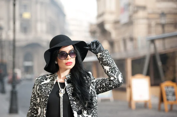 Jovem atraente em um tiro de moda de inverno. Bela menina na moda em preto posando na avenida. Morena elegante com chapéu, óculos de sol e luvas no cenário urbano . — Fotografia de Stock